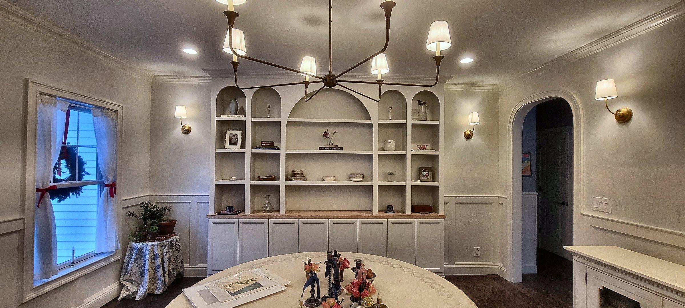 Arched built-in bookcases on either side of a dining room, with wainscot below and a chandelier between them.