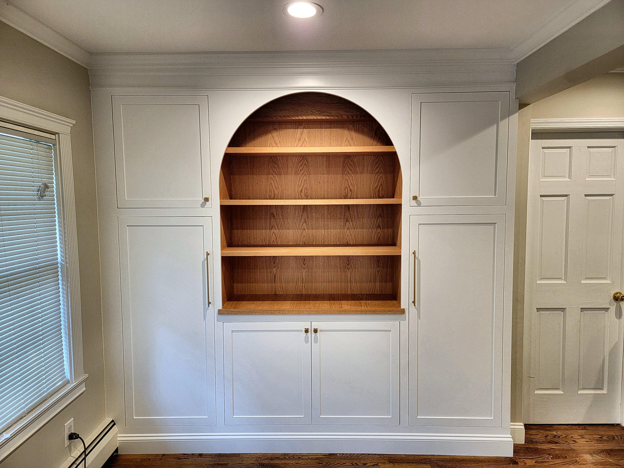 A white arched built-in with oak shelves, closed storage below, and a round window in the wall beside it.